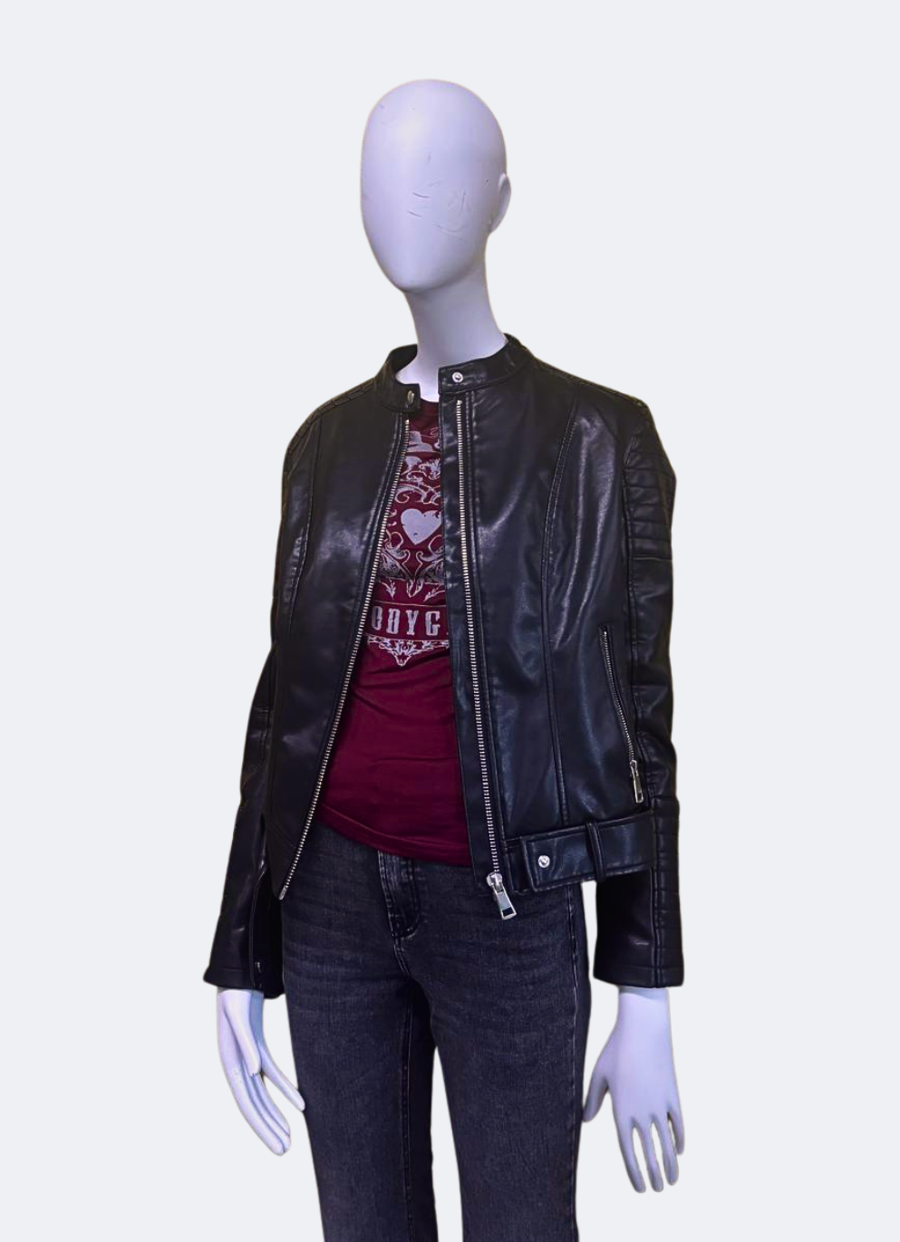 Menequí posando de manera lateral con una chaqueta de cuero mujer negra, de la marca Urban Mirage, combinada con camiseta roja y jeans. El estilo es casual con cremallera y detalles modernos plateados. Ideal para outfits streetwear.